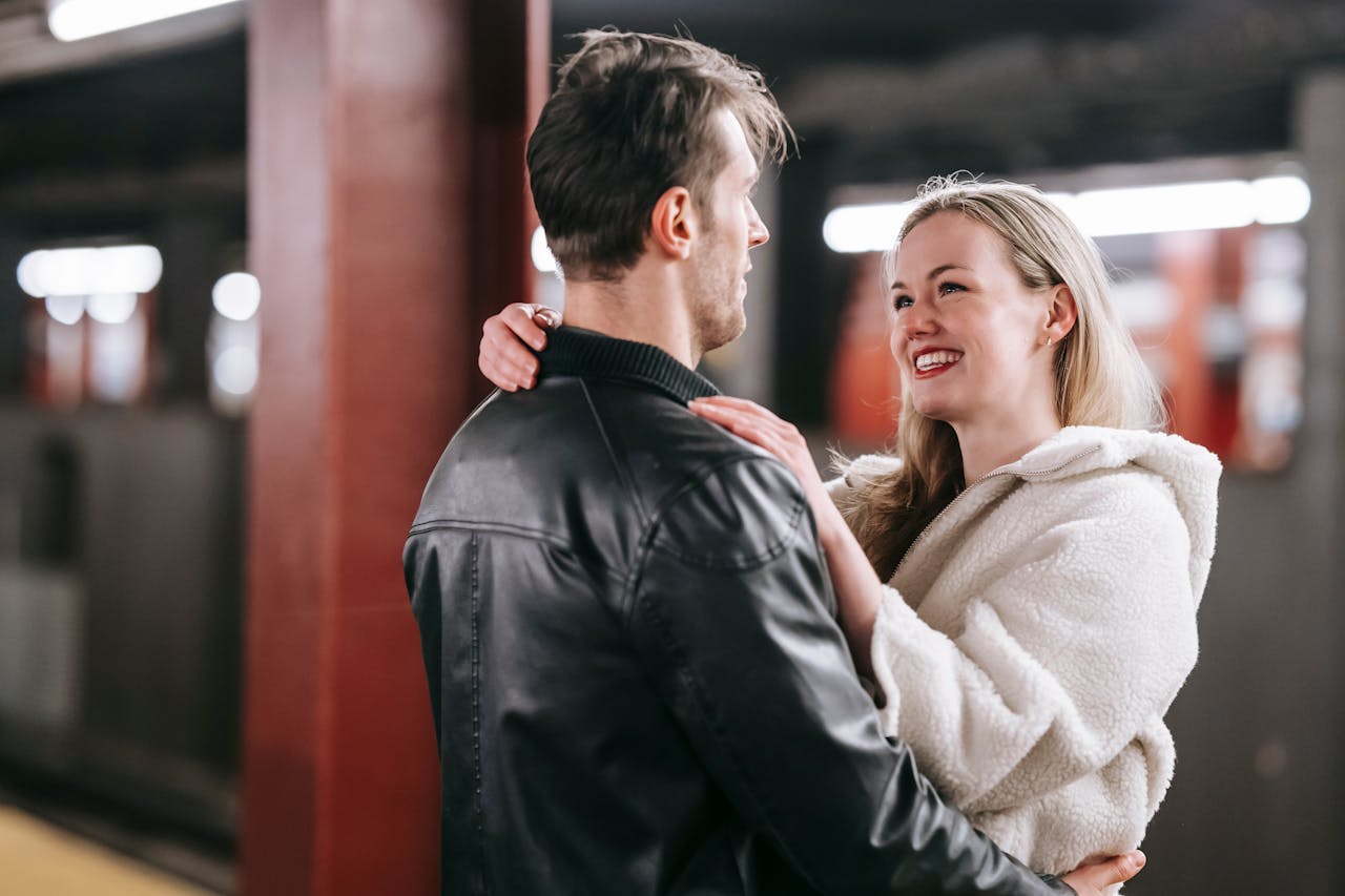 Smiling couple looking at each other and embracing while standing on platform of metro with train on blurred background during date